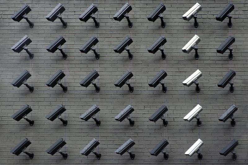 Grid of surveillance cameras mounted on a grey brick wall, mostly black with a few white ones, all pointing in the same direction