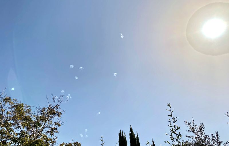 Iron Dome missile interceptions visible as white smoke puffs scattered across a blue sky over northern Israel, with trees silhouetted in the foreground