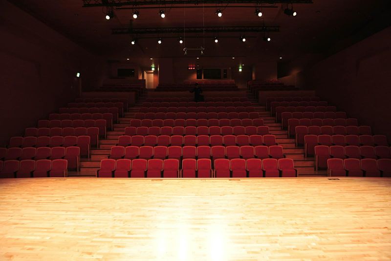 An illuminated empty theater stage with rows of red seats stretching into darkness, viewed from the performer's perspective