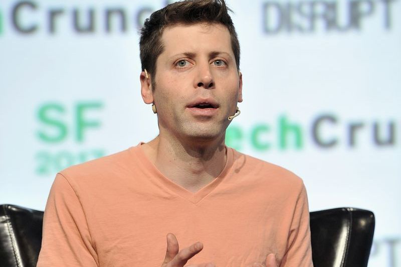 Sam Altman speaking on stage at TechCrunch Disrupt SF 2017, gesturing with both hands while seated in a black leather chair