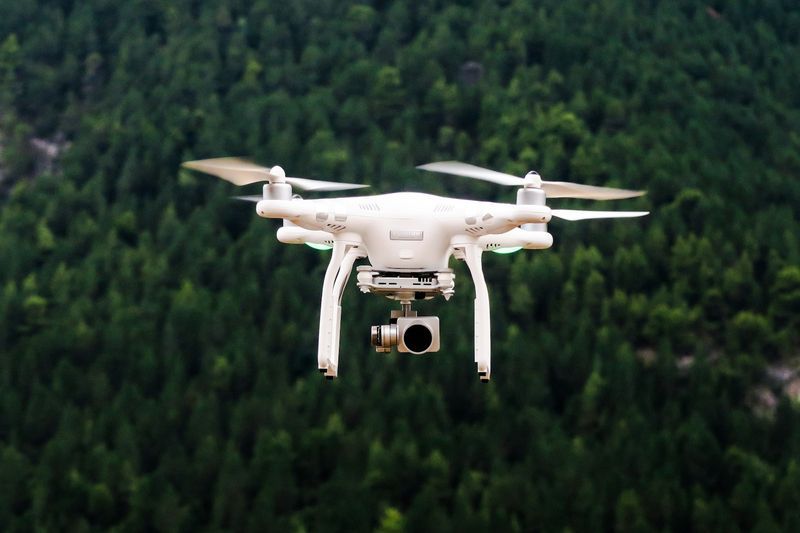 White quadcopter drone with camera hovering in flight above a forested hillside