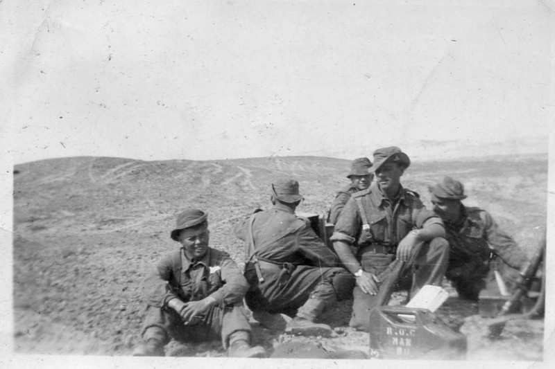 A group of soldiers in a black and white photograph from World War II, outdoors on rugged terrain.