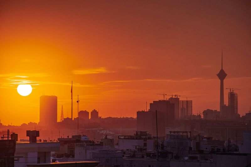 Tehran skyline silhouetted against a deep orange sunset, with the Milad Tower rising on the right and construction cranes dotting the hazy cityscape
