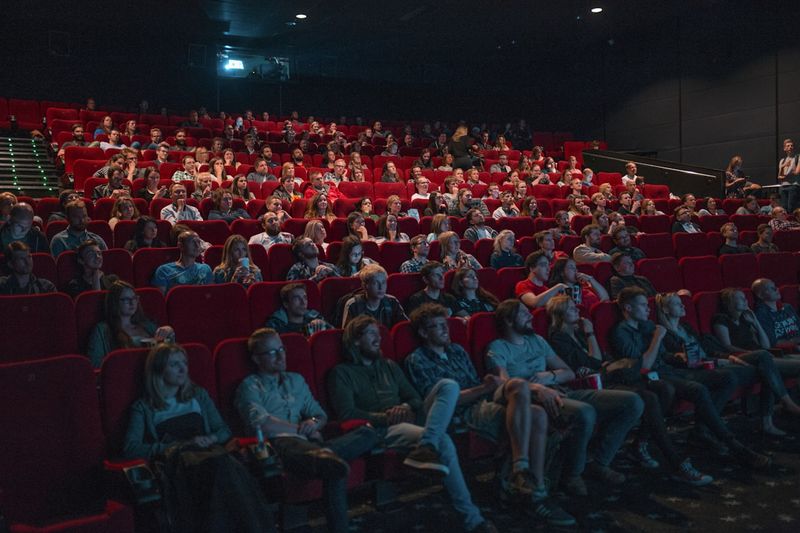 A packed movie theater audience watching a film, with rows of red seats filled with engaged viewers in a dimly lit cinema