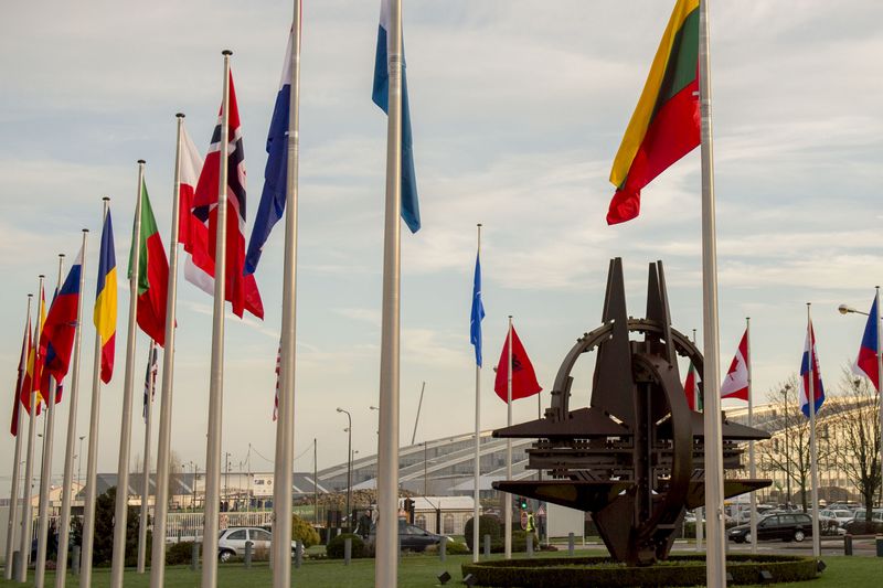 Flags of NATO member countries waving on flagpoles at the entrance of NATO headquarters in Brussels, with the NATO Star sculpture visible in the center