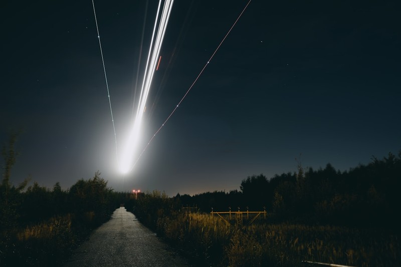 Long-exposure photograph of bright light trails streaking across a dark night sky above a rural road, evoking the paths of drones and missiles during a nighttime aerial attack