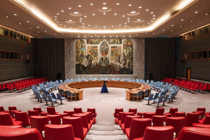 The United Nations Security Council chamber in New York, showing the curved delegates' table, blue and red seating, and the iconic Per Krohg mural on the back wall