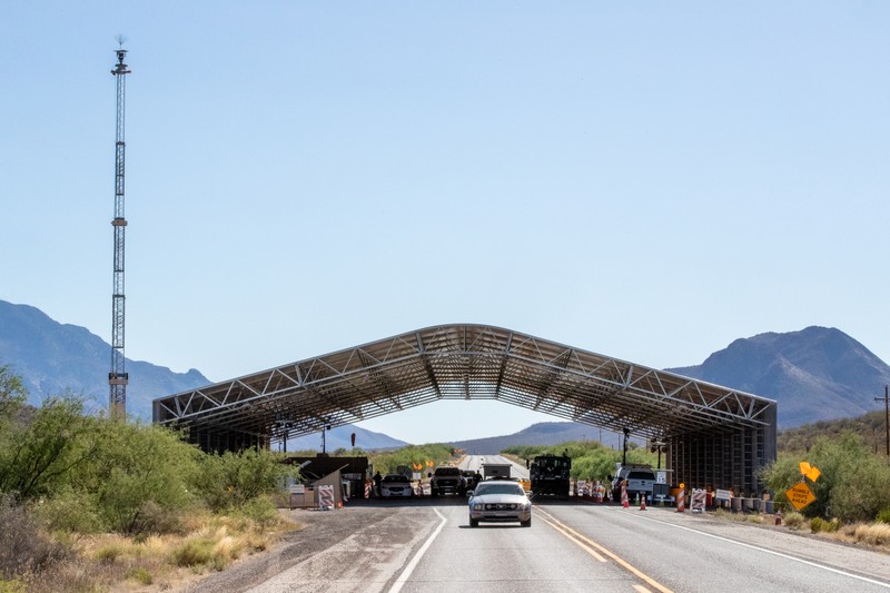 A U.S. Border Patrol checkpoint station on a desert highway, with vehicles approaching a large covered inspection canopy and mountains in the background