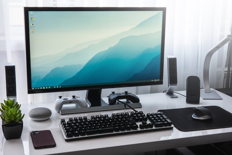 A Windows desktop computer setup with monitor displaying a mountain landscape wallpaper, mechanical keyboard, mouse, and speakers on a white desk