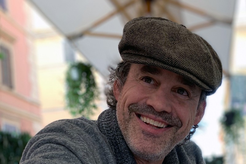 Nicholas Brendon smiling while wearing a flat cap at an outdoor café in Italy