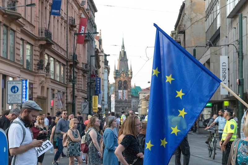 Crowd marching through a Prague street with a European Union flag waving prominently, historic architecture and church spire visible in the background