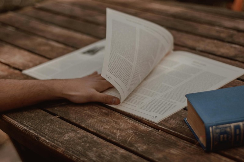 A person's hand turning the pages of an open book on a wooden table, examining the printed text closely