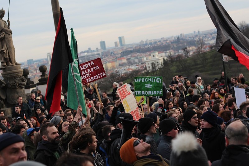 Large crowd of demonstrators holding flags and protest signs gathered near Prague Castle with the city skyline in the background