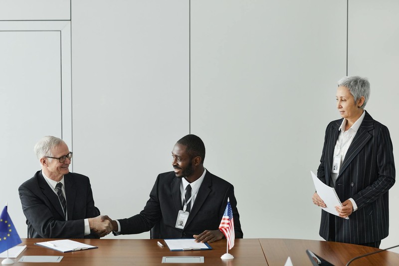 Diplomats shaking hands across a conference table with American and European Union flags during formal negotiations