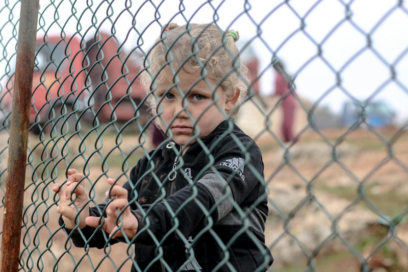 A young child stands behind a chain-link fence, gripping the wire and looking out with a solemn expression