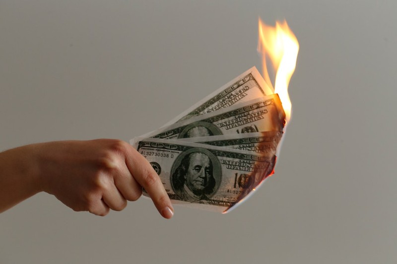A hand holding several hundred-dollar bills engulfed in flames against a plain background