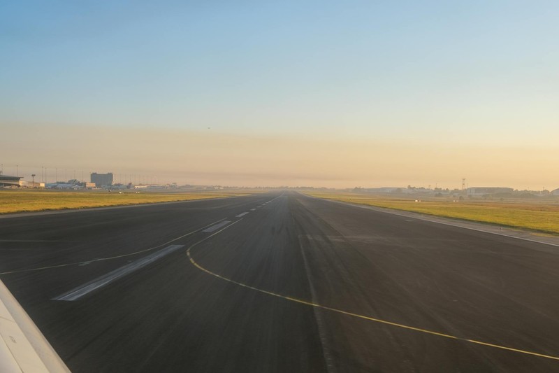 An empty airport runway at sunrise, showcasing a clear sky and distant airport buildings.