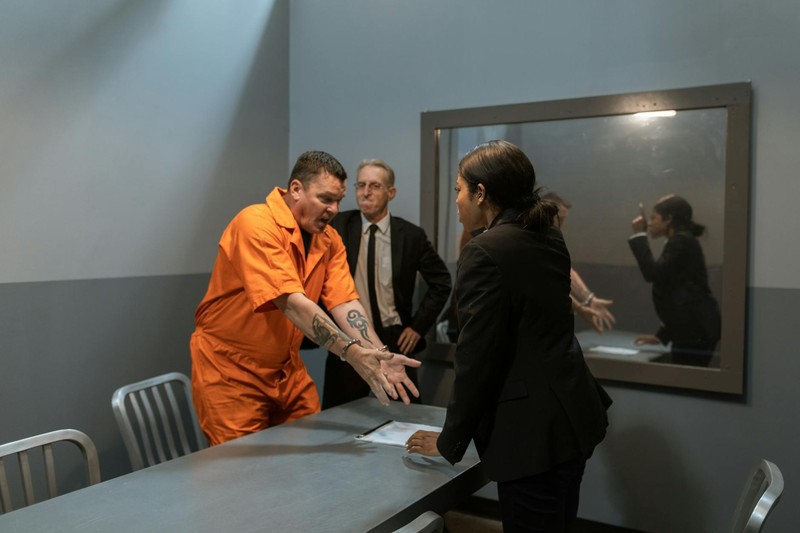 A prisoner in an orange jumpsuit speaks with officers in an interrogation room