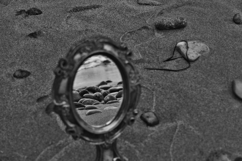 An ornate mirror standing in dark sand, reflecting a curated arrangement of stones while the surrounding beach shows a different reality