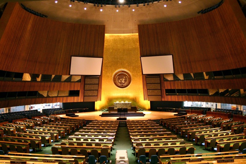 Interior view of the United Nations General Assembly hall with curved seating arrangement and UN emblem prominently displayed at the front