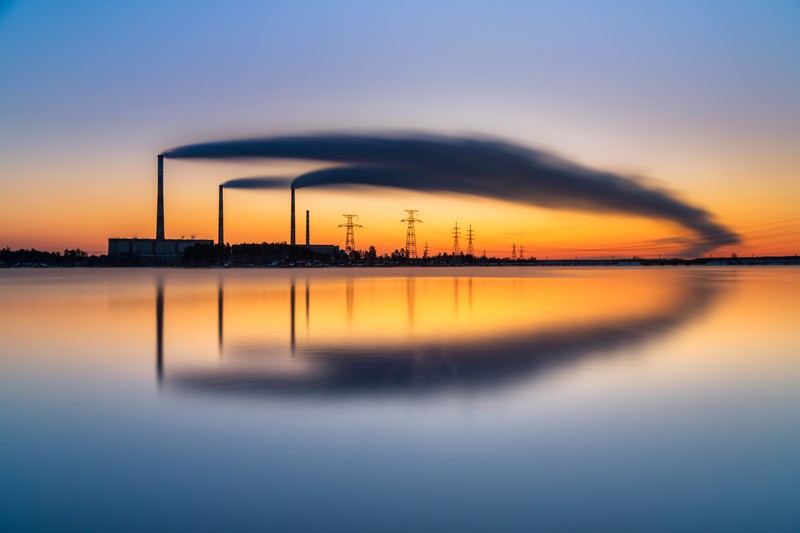 Industrial smokestacks emitting dark smoke plumes at sunrise over a calm reservoir with perfect reflections