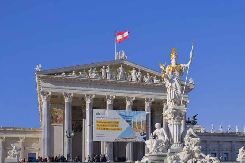 The Austrian Parliament Building in Vienna with a golden statue in the foreground and the Austrian flag flying atop