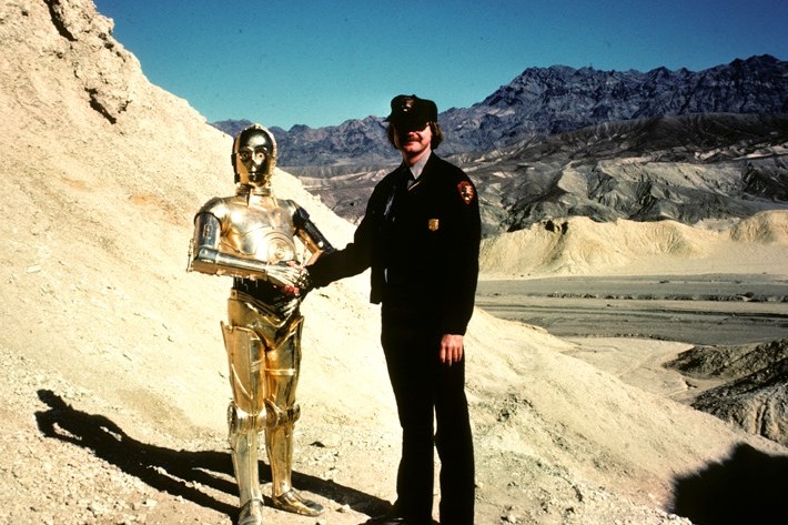 C-3PO droid shakes hands with a uniformed person in a desert landscape during Star Wars filming