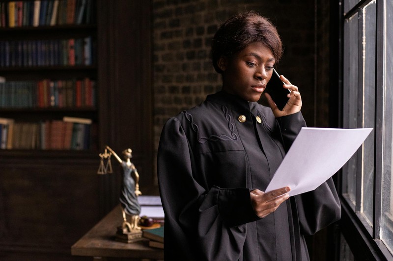 A judge in a black robe with gold buttons holds a document while speaking on a phone, with a Lady Justice statue visible in the background