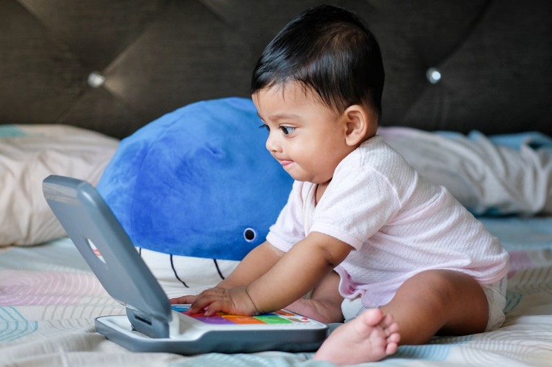 A young toddler sitting on a bed, focused on a colorful toy laptop with both hands on the keyboard