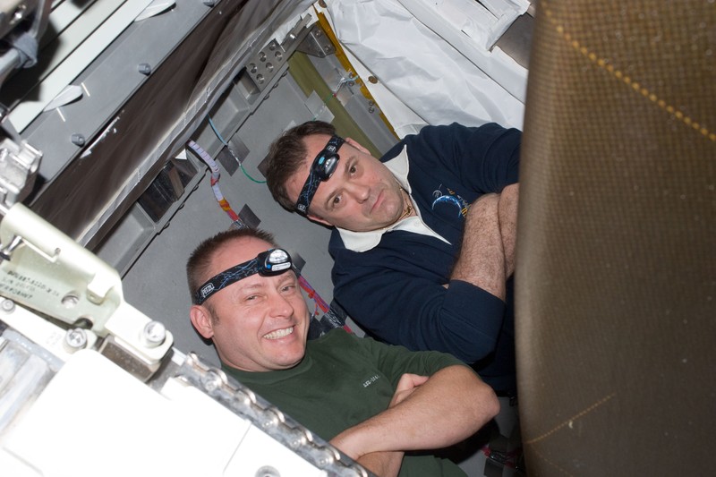 Two astronauts work inside the International Space Station, wearing headlamps in the dimly lit interior