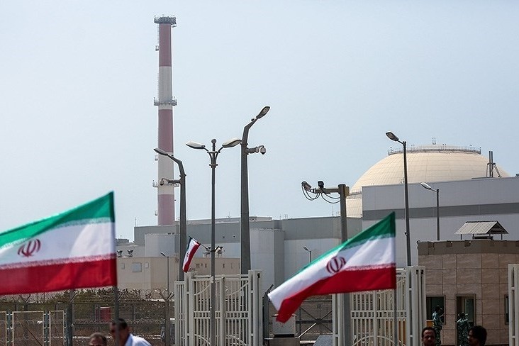 Iranian flags frame the entrance to the Bushehr Nuclear Power Plant, with the reactor containment dome and smokestack visible in the background.