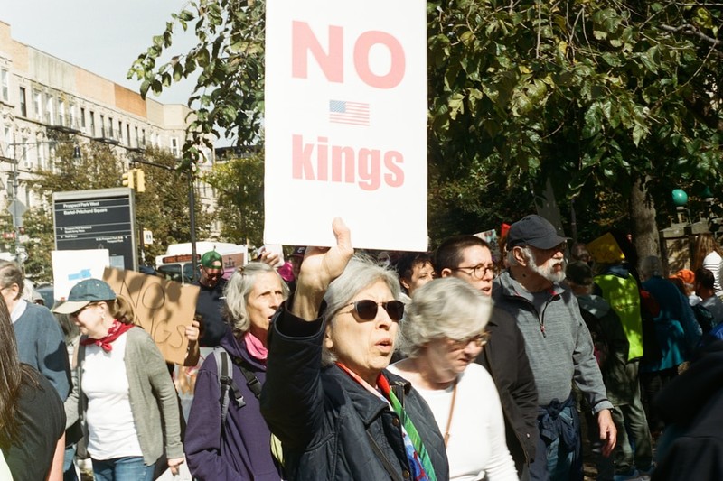 A protester at a No Kings demonstration holds a white sign reading 'NO KINGS' with a small American flag, surrounded by fellow demonstrators in an urban setting.
