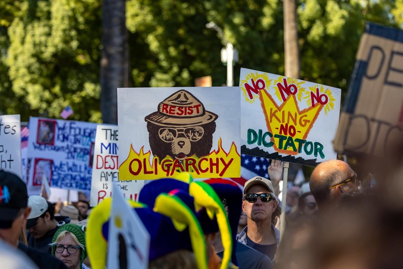 Protesters holding anti-oligarchy and No King No Dictator signs at a crowded outdoor demonstration