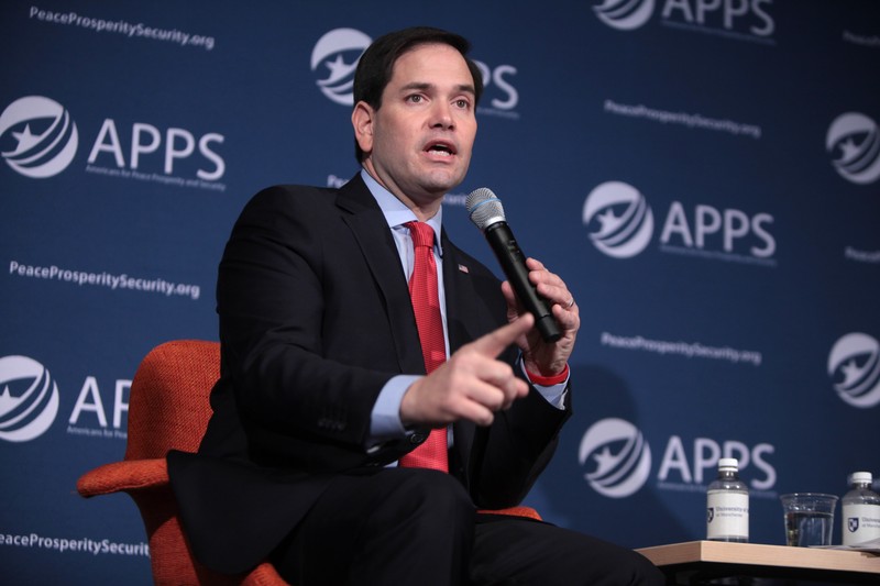 Marco Rubio speaking at a policy forum with a microphone, wearing a dark suit and red tie