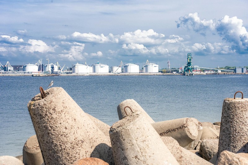 Industrial port with large cylindrical oil storage tanks, concrete breakwaters, and harbor infrastructure under a partly cloudy sky