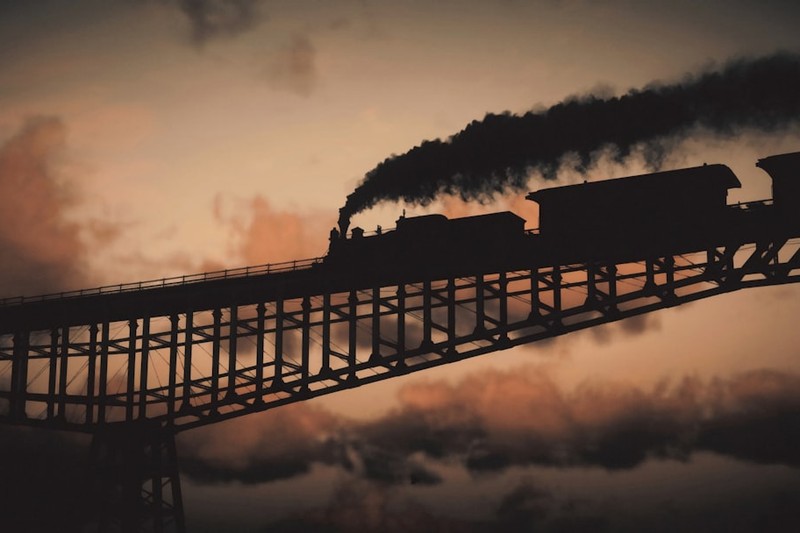 A steam train crosses a steel truss bridge at sunset in Red Dead Redemption 2