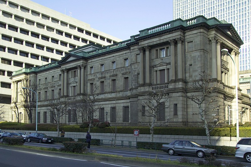 The Bank of Japan headquarters building in Tokyo's Nihonbashi district, with modern skyscrapers rising behind its neoclassical stone facade and green copper roof