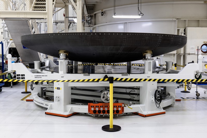 A large circular spacecraft heat shield mounted on a work stand inside a NASA facility at Kennedy Space Center