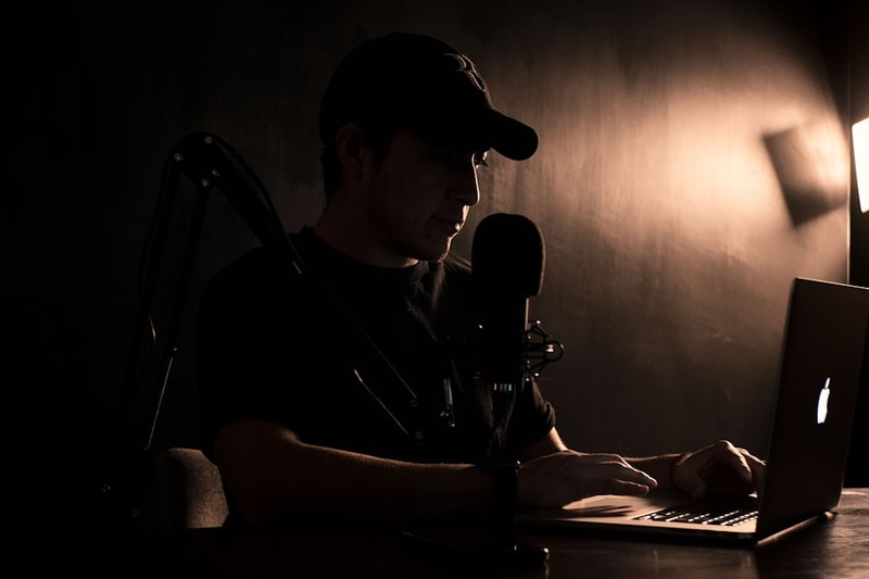A man wearing a dark baseball cap records a podcast at a desk with a microphone and laptop in a dimly lit room
