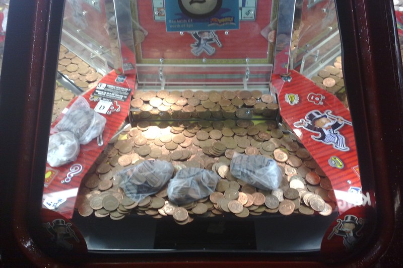 A coin pusher arcade machine filled with pennies and wrapped prizes, viewed through its glass front.