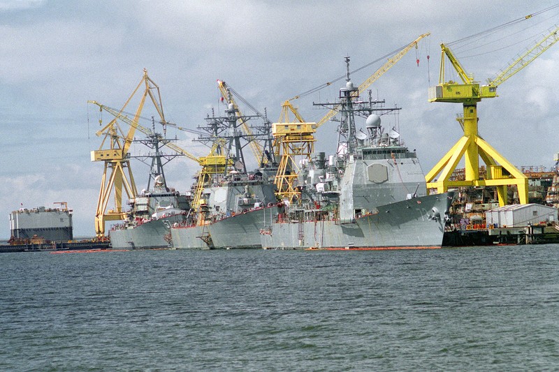 U.S. Navy warships docked at a shipyard with yellow cranes in the background under overcast skies