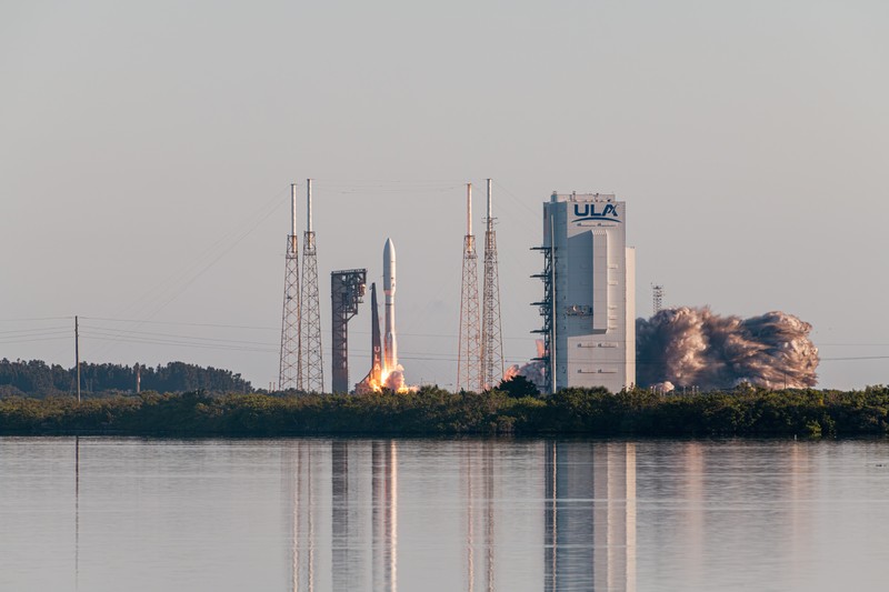 An Atlas V rocket ignites on the launch pad at Cape Canaveral, carrying Amazon's Kuiper 3 satellite mission, with its reflection visible in calm water