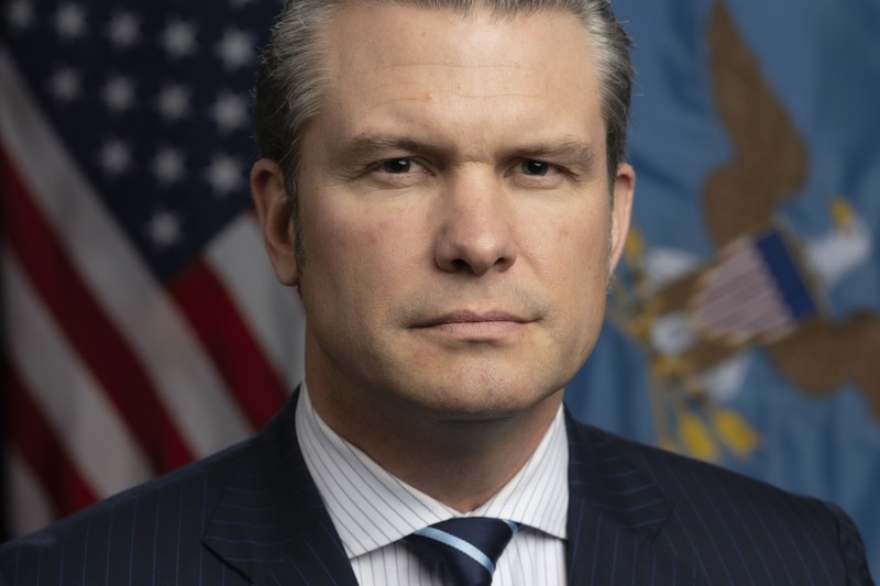 Official portrait of Secretary of Defense Pete Hegseth in a dark suit with U.S. flags in the background