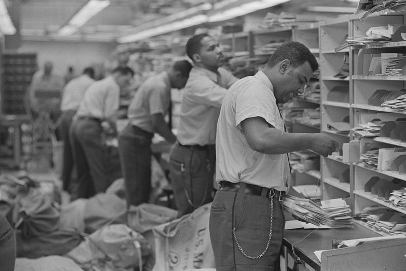 Black-and-white photograph of postal workers sorting mail in a busy facility