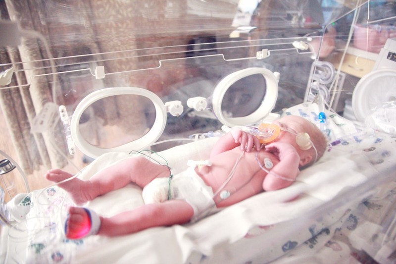 A premature baby lying in a clear isolette incubator in a neonatal intensive care unit, connected to oxygen and monitoring equipment