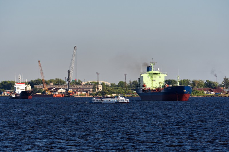 A large oil tanker navigates a wide river past industrial port infrastructure under an overcast sky