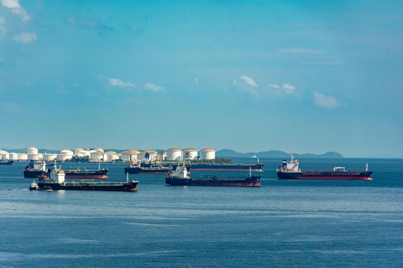 Cargo ships and oil storage tanks along a coastal waterway under a clear blue sky