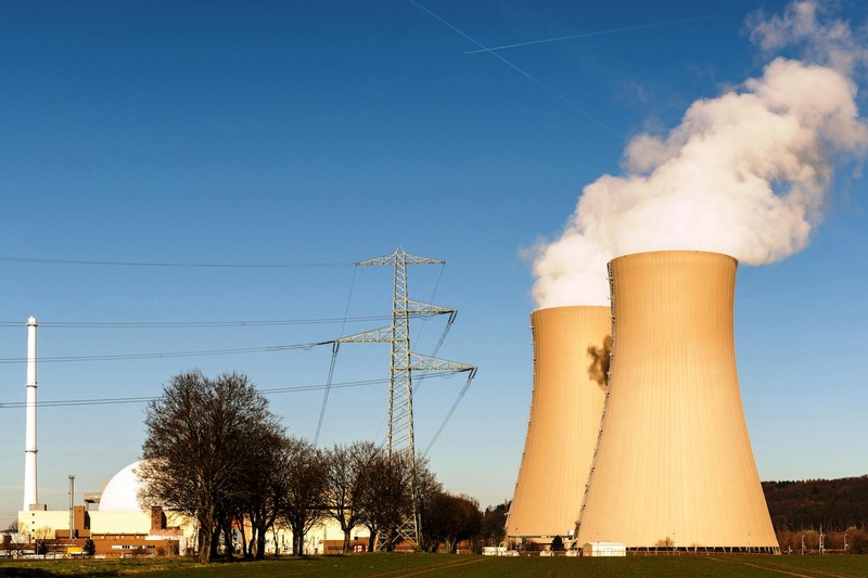 A nuclear power plant with cooling towers releasing steam, a domed reactor containment building, and high-voltage transmission towers under a clear blue sky.