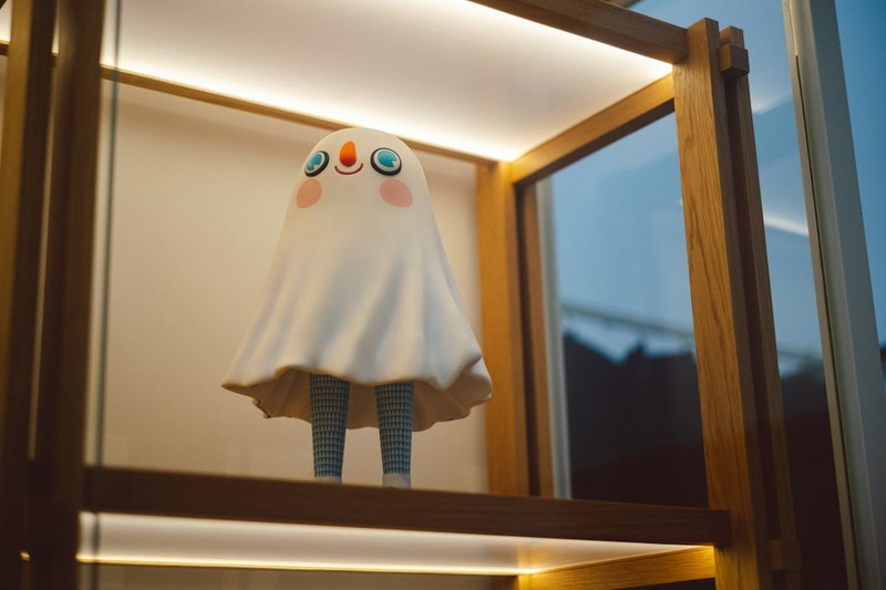 A small stuffed ghost figure with round eyes and pink cheeks stands alone on a wooden display shelf