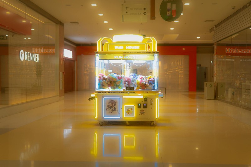 A brightly lit claw machine filled with colorful plush toys sits alone in a deserted shopping mall corridor, its neon glow reflected on the polished floor.